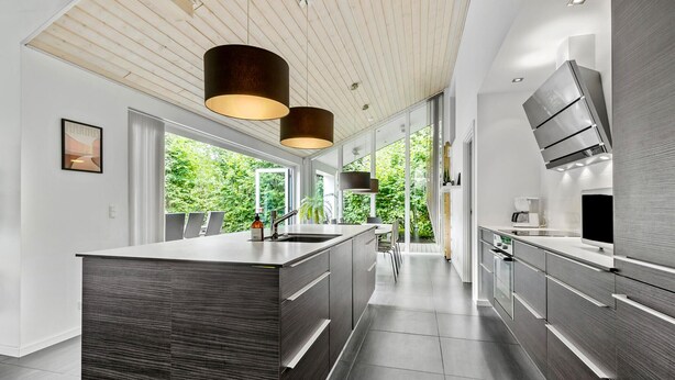 A modern kitchen with a central island, pendant lighting, and large windows offering a view of greenery.
