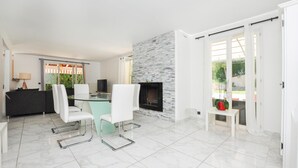 Ein modernes Wohnzimmer mit einem Glastisch, weißen Stühlen, einem schwarzen Sofa, einem Kamin und Blick auf Grünflächen.
