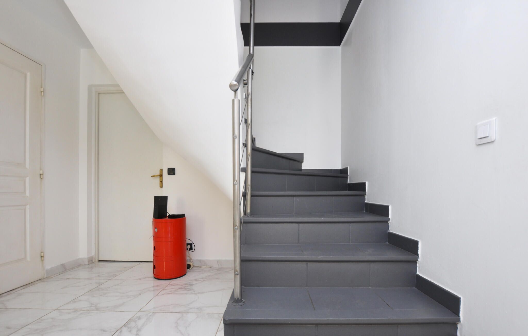 Un escalier avec une rampe en métal, des murs blancs et un objet cylindrique rouge posé sur le sol.