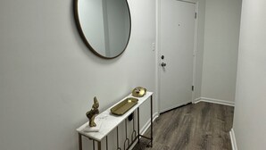A hallway with a white door, a round mirror, a marble countertop with a gold tray, and a metal console with a statue.