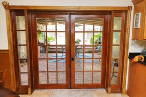 Une porte double en bois, dotée de panneaux de verre, donne sur un patio extérieur avec un sol en briques et du mobilier de jardin.