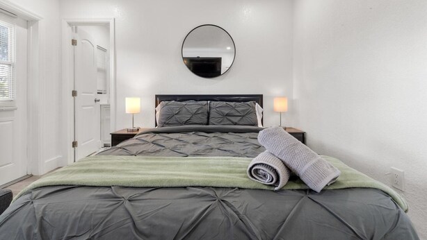 Quarto com cama, espelho redondo, duas luminárias de cabeceira e uma porta.