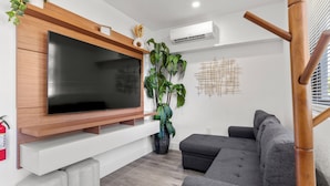Uma sala de estar moderna com televisão de tela plana presa a uma prateleira de madeira, um sofá cinza, uma planta grande em vaso e um aparelho de ar condicionado.