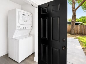 Une buanderie avec un lave-linge et un sèche-linge blancs, une porte noire et une vue sur un jardin avec un arbre et une clôture.