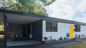 Uma casa moderna térrea com varanda coberta, porta amarela e um pequeno jardim com diversas plantas.