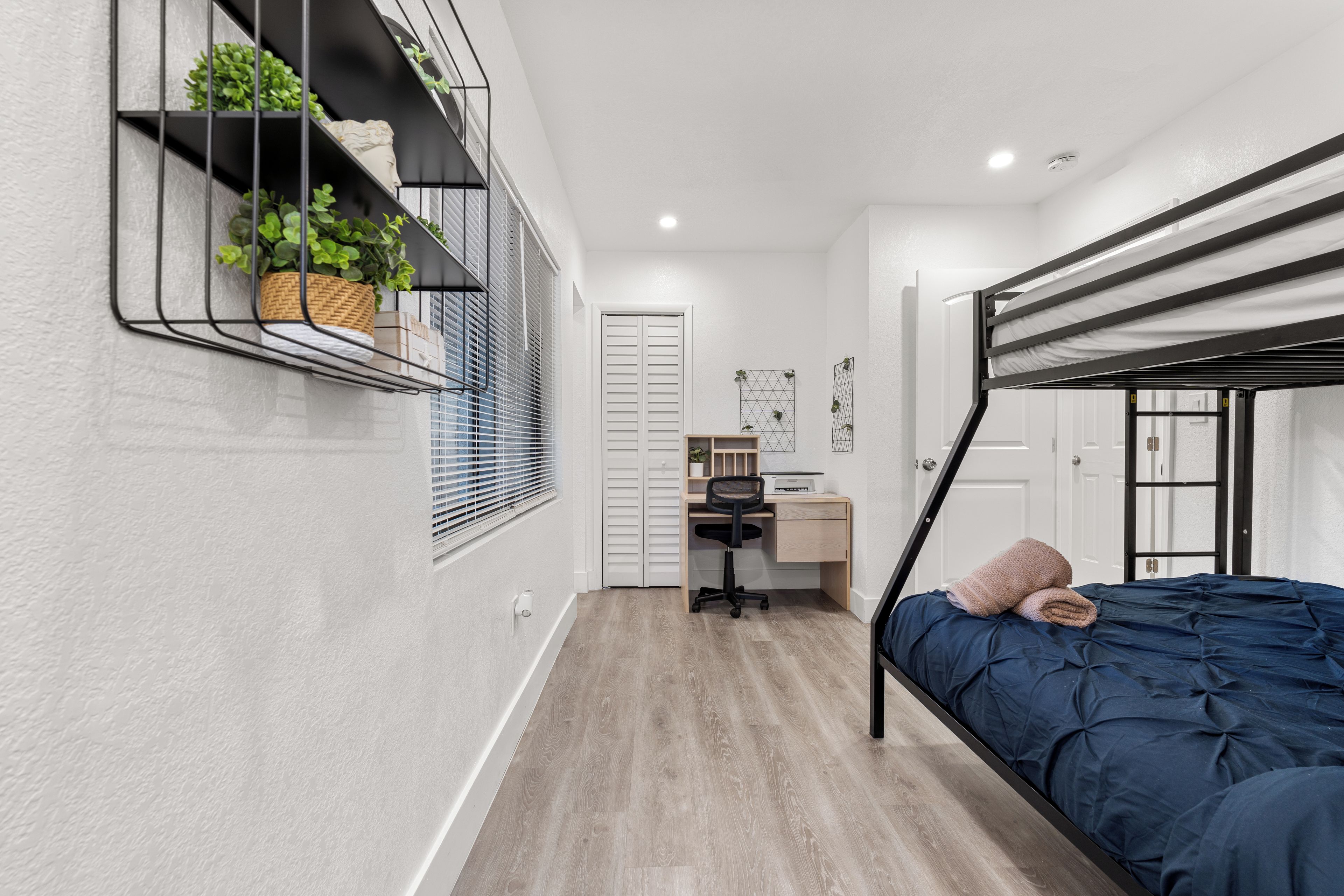 Une chambre à coucher de taille réduite, avec un lit superposé, un bureau avec une chaise et une étagère avec des plantes.