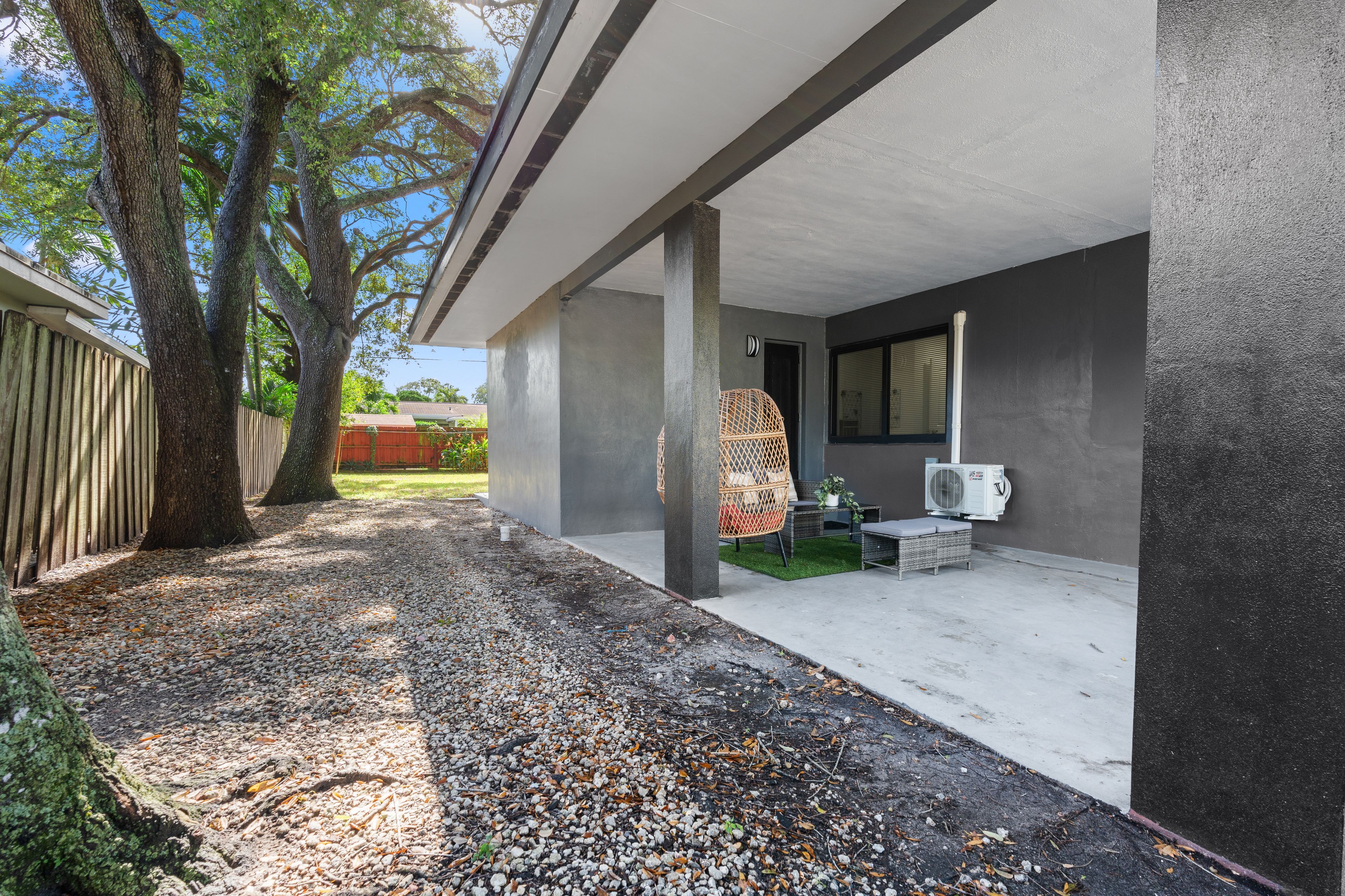 A covered outdoor area with a concrete floor, a wicker chair, and a small table with a potted plant.