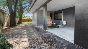 Uma área externa coberta com piso de concreto, uma cadeira de vime e uma pequena mesa com um vaso de planta.