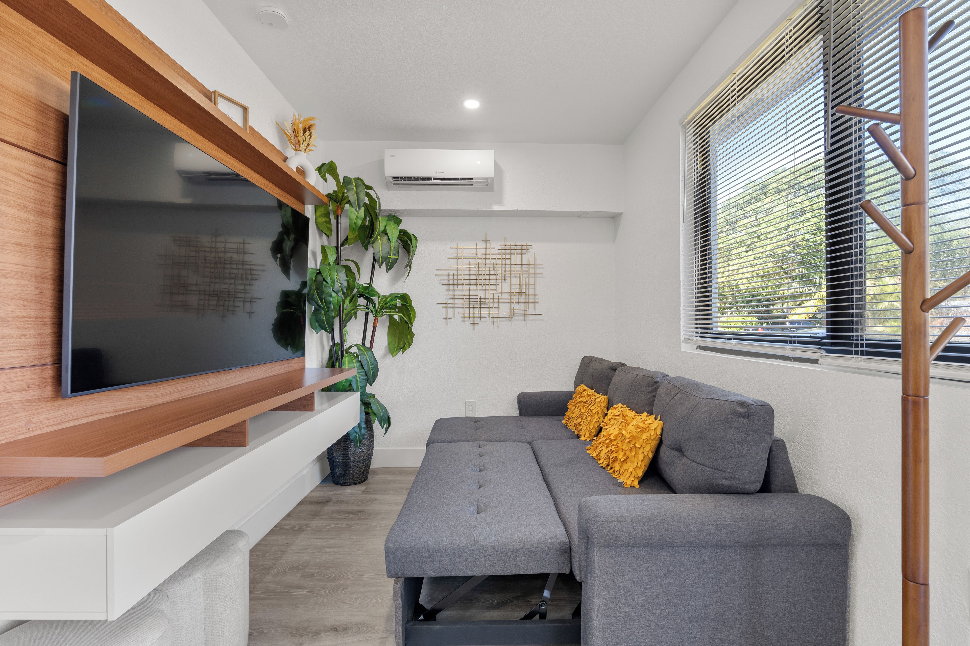 Uma sala de estar moderna com uma TV de tela plana presa em uma prateleira de madeira, um sofá cinza com almofadas amarelas, uma planta grande em vaso e uma janela com persianas.