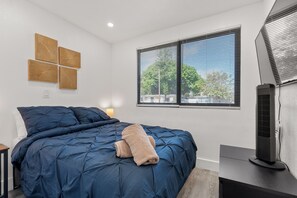 A bedroom with a large bed, a bedside table, a lamp, a window with blinds, and a wall-mounted air conditioning unit.