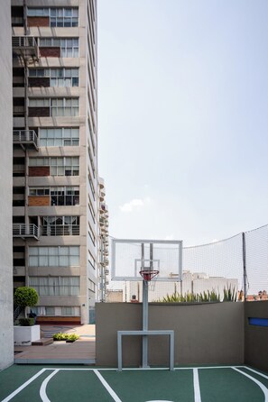 An outdoor basketball court with a net, surrounded by a fence, and adjacent to a high-rise building.