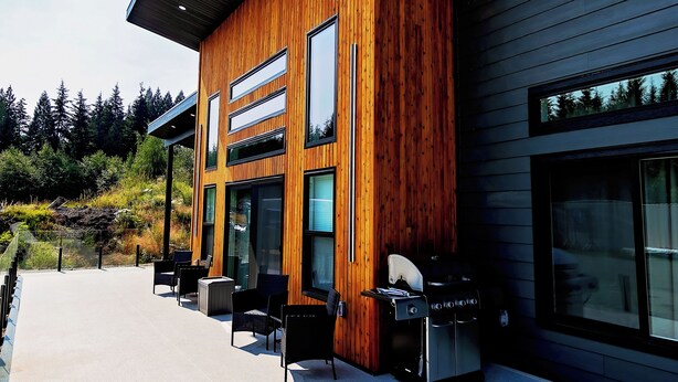 Un bâtiment moderne doté d’une façade en bois, de grandes fenêtres et d’un patio aménagé avec des espaces de détente en plein air et un barbecue.