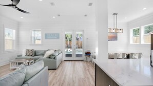 Un vaste salon avec un canapé gris, une table basse et un coin repas comprenant une longue table et des chaises. Un panneau sur le mur porte l’inscription « PLAGE ».