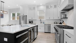 Une cuisine moderne avec des meubles blancs, des appareils en acier inoxydable, un îlot central et une fenêtre équipée de stores.