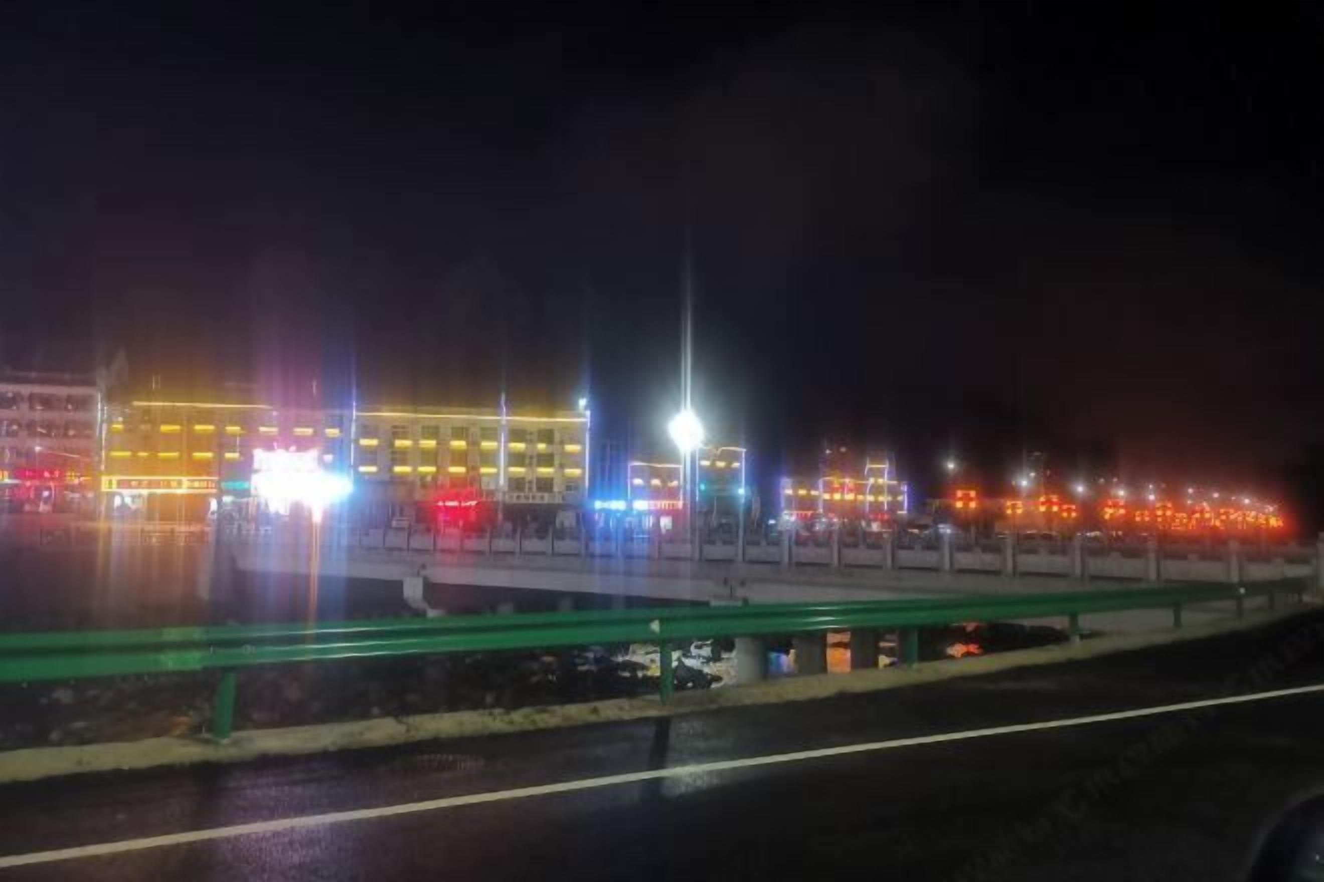 Vista noturna de um edifício iluminado, com vários andares e uma ponte verde em primeiro plano.