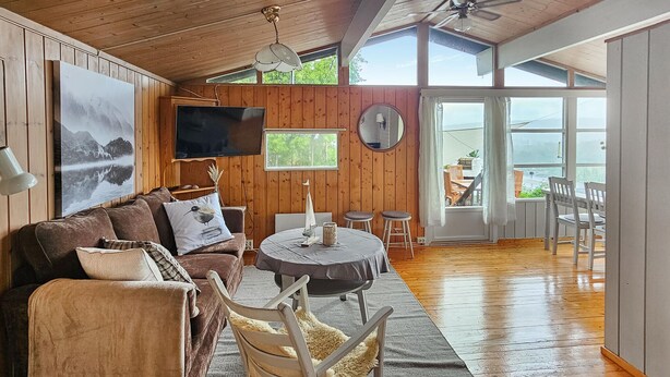 Una sala de estar acogedora con techo de madera, televisor grande, mesa redonda y vistas al exterior a través de amplios ventanales.