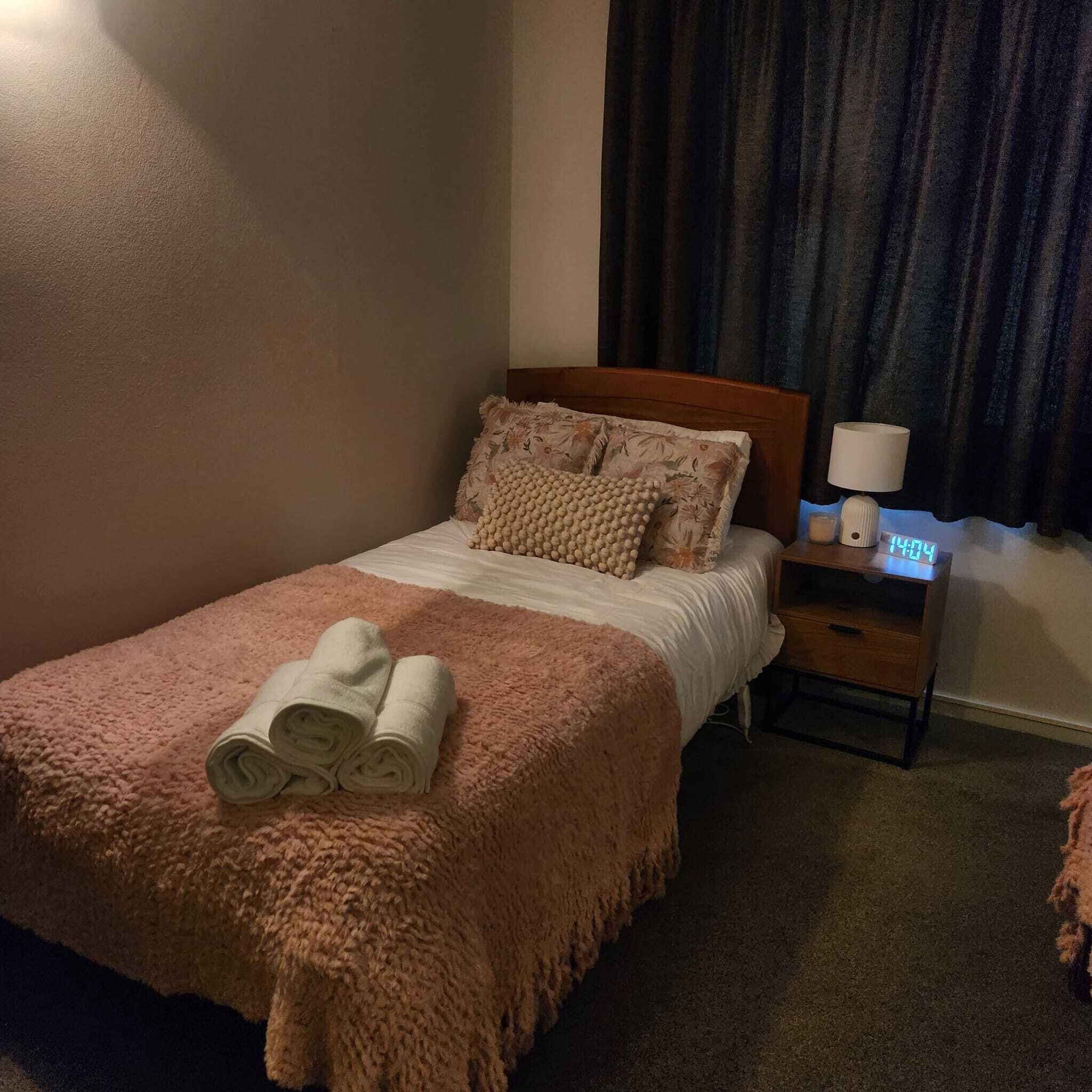 En pænt redt seng med et rosa tæppe og sammenrullede håndklæder, et trænatbord med en lampe og et digitalt ur, samt en mørk gardin i baggrunden.