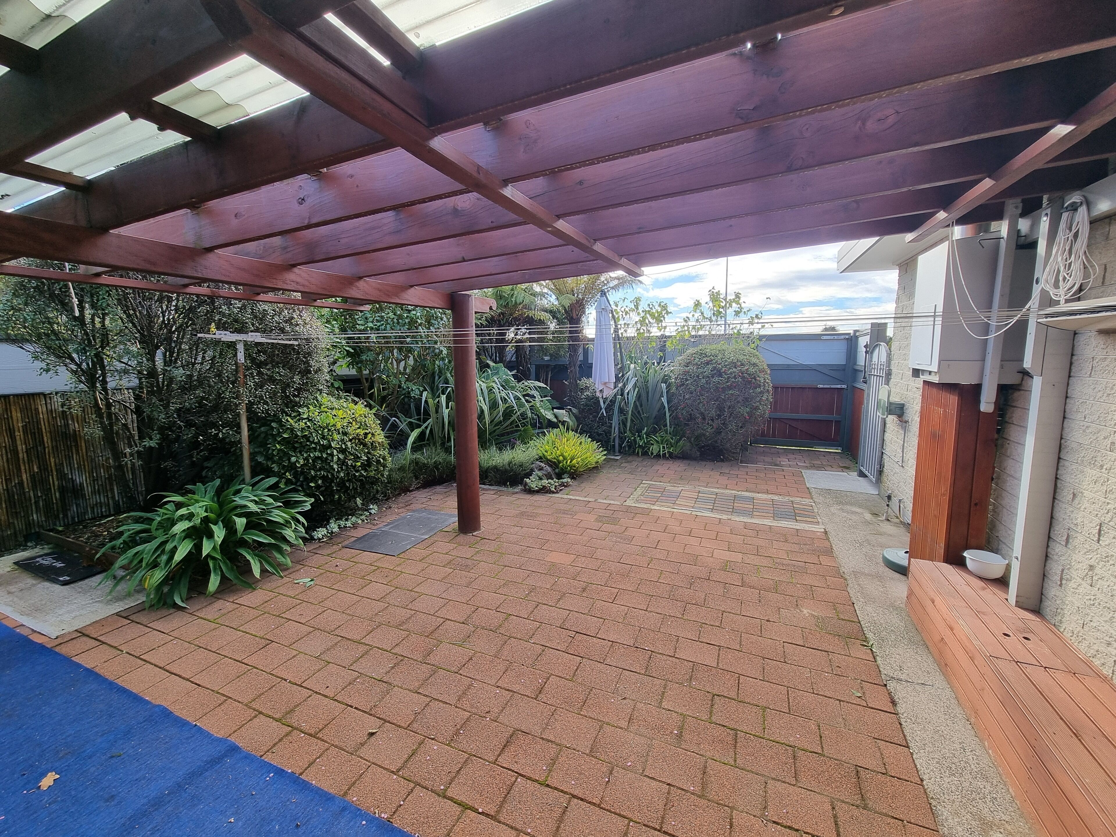 A covered patio area with brick flooring, a wooden pergola, and various plants.