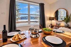 A dining table set with plates, glasses, a bottle, and a bowl of salad, with a view of a snowy landscape through large windows.