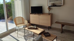A modern living room with a large window, a television on a wooden cabinet, a wicker chair, a small table with decorative items, and a bench. - House with covered swimming pool and pétanque court (Lanchéres)