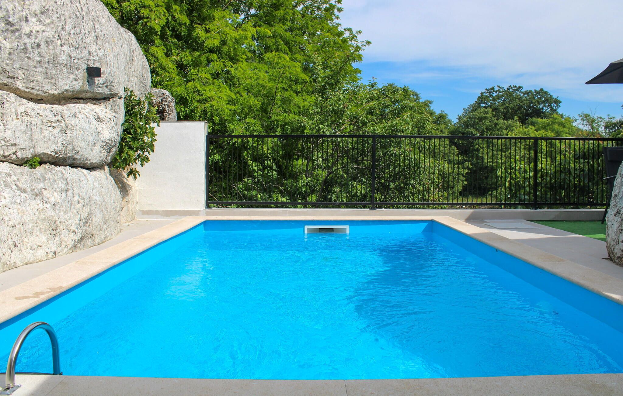 A rectangular swimming pool with clear blue water, surrounded by a black metal fence, and a large rock structure to the left.