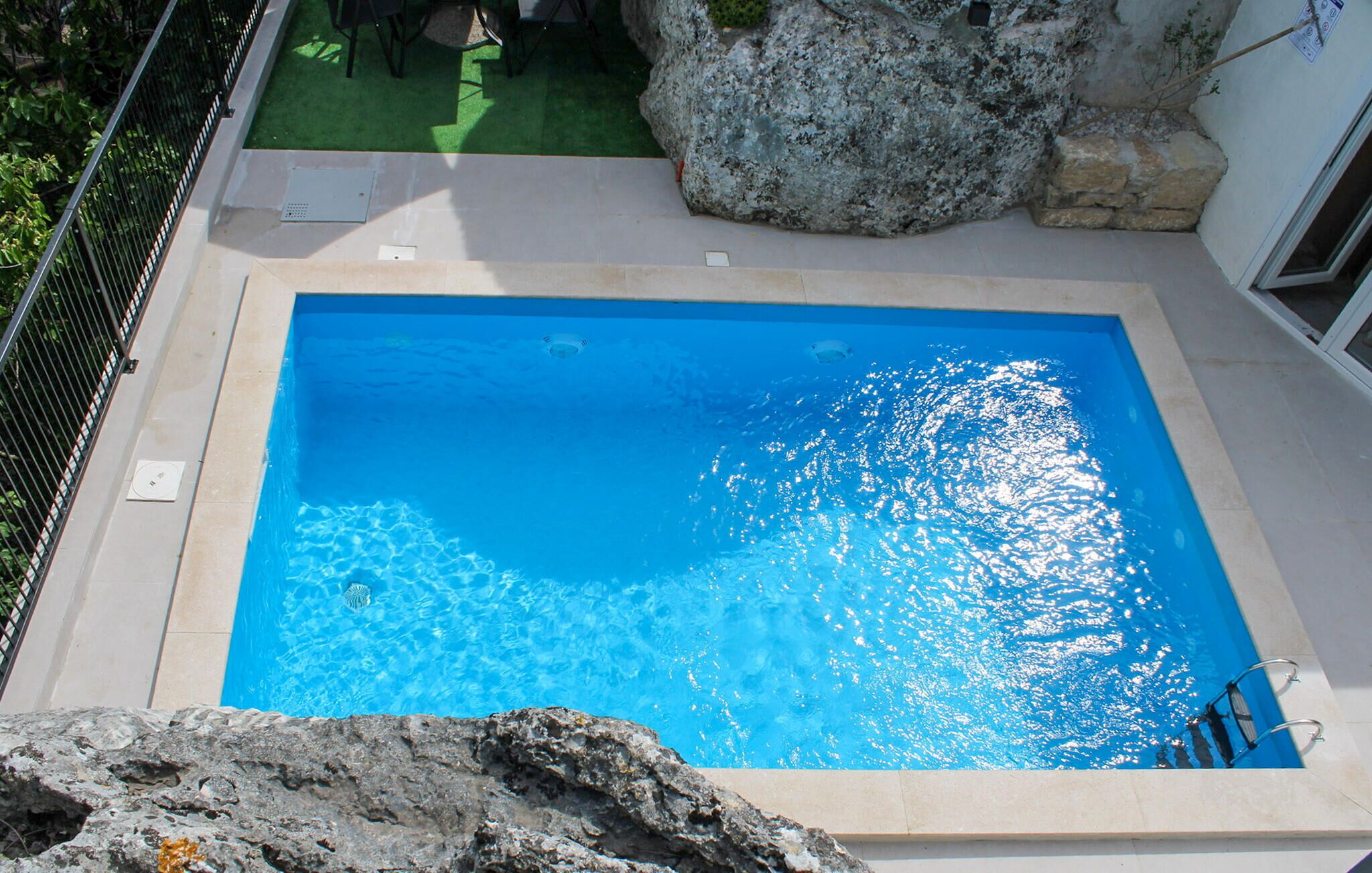 A rectangular swimming pool with clear blue water, surrounded by a stone wall and a metal fence.
