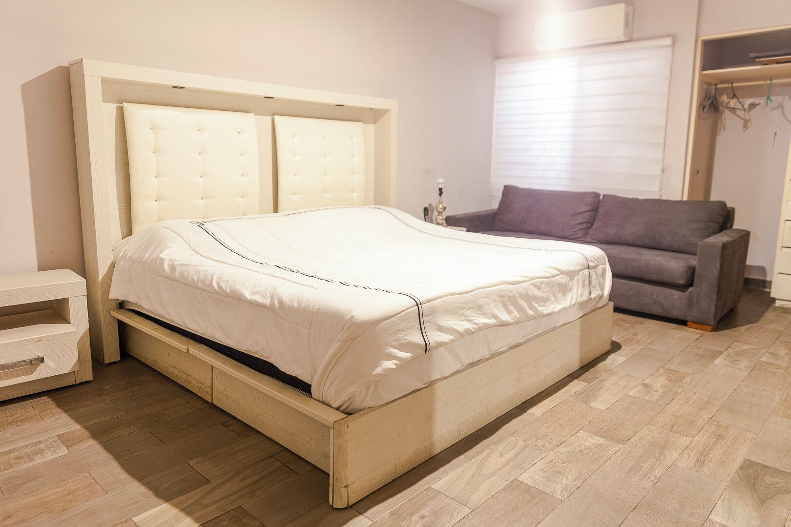 A bedroom with a bed, a bedside table, a grey sofa, and a window with blinds.