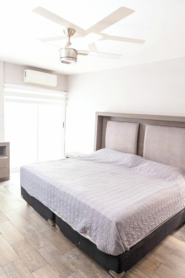 A bedroom with a bed, a ceiling fan, an air conditioning unit, and a wooden floor.