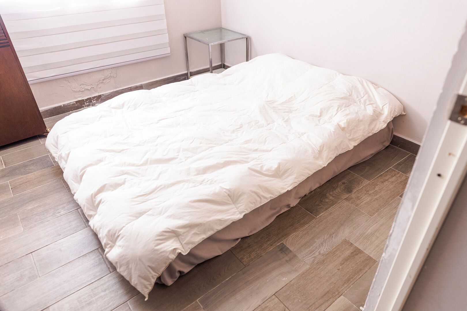 A small, single-bed room with a wooden floor, a bedside table, and a window with blinds.