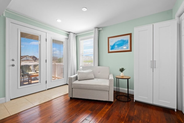 Une pièce avec un canapé gris, une petite table ronde ornée d’une plante, une armoire blanche et une porte donnant sur un balcon aménagé avec du mobilier d’extérieur.