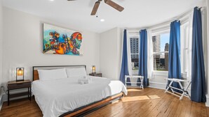 Camera da letto con un letto, una ventola a soffitto, un quadro, una finestra con tende e vista sugli edifici.