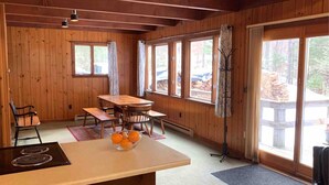 L’intérieur d’un chalet en bois comprend une table à manger, des chaises, un poêle et une grande fenêtre donnant sur l’extérieur.