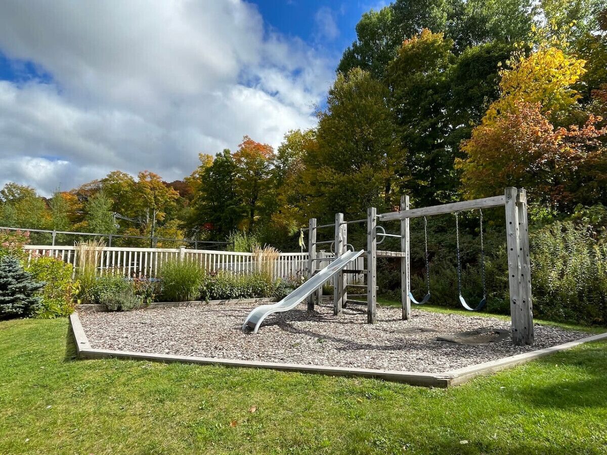A playground with a slide and swings, surrounded by a wooden fence and a gravel area.
