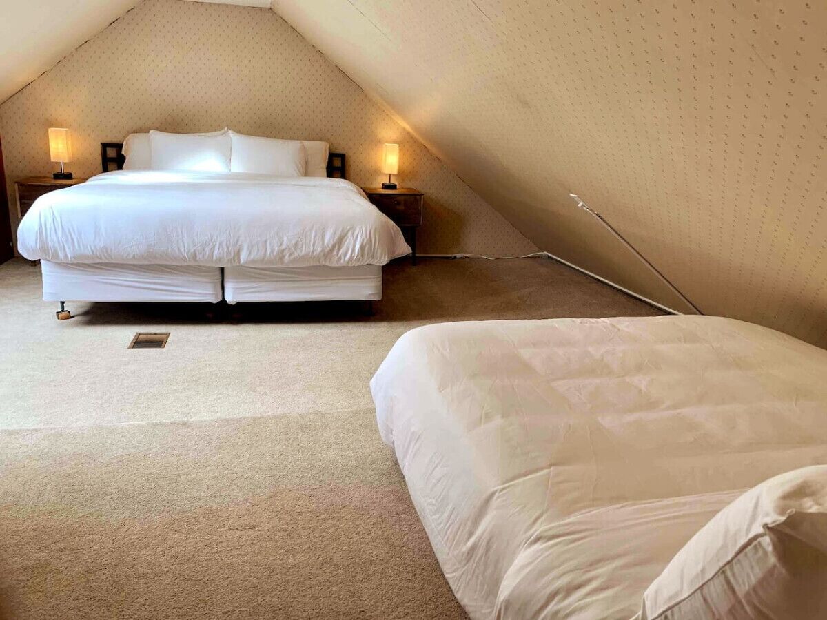 Une chambre à coucher avec un grand lit et un plus petit lit, tous deux dotés de draps blancs, située dans une pièce mansardée avec des plafonds inclinés et deux lampes de chevet.