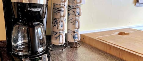 A coffee maker, a stack of mugs, and a wooden cutting board on a kitchen counter.