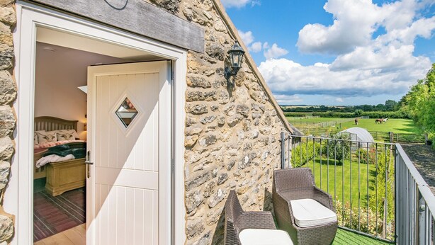 A stone building with a bedroom visible through an open door, a balcony with wicker furniture, and a view of a green field with horses. - 1 bedroom accommodation in Pickering (Pickering)