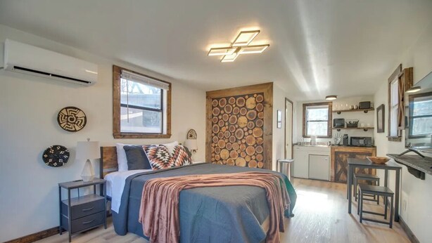 A modern bedroom with a large bed, a small bedside table, a wall-mounted air conditioner, and a kitchen area in the background.