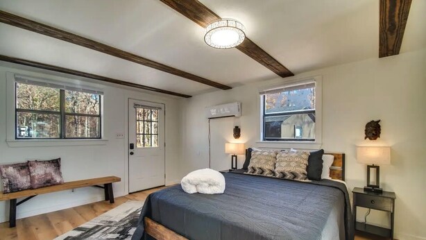 Une chambre avec un grand lit, un banc, deux fenêtres, un ventilateur de plafond et un climatiseur fixé au mur.
