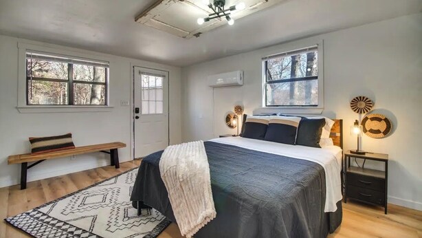 Une chambre avec un grand lit, deux fenêtres, un banc, une table de chevet avec une lampe et un ventilateur de plafond.