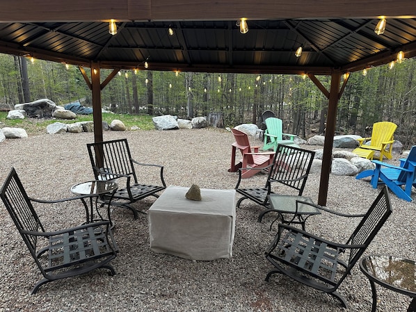 Ein Sitzbereich im Freien mit Metallstühlen und einem Tisch, umgeben von Kieselsteinen und Felsen, unter einer Holzpergola mit Lichterketten.