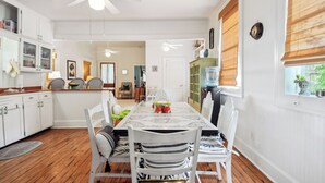 Un coin repas avec une table dressée pour quatre personnes, des chaises blanches, un ventilateur de plafond et une fenêtre avec des stores.