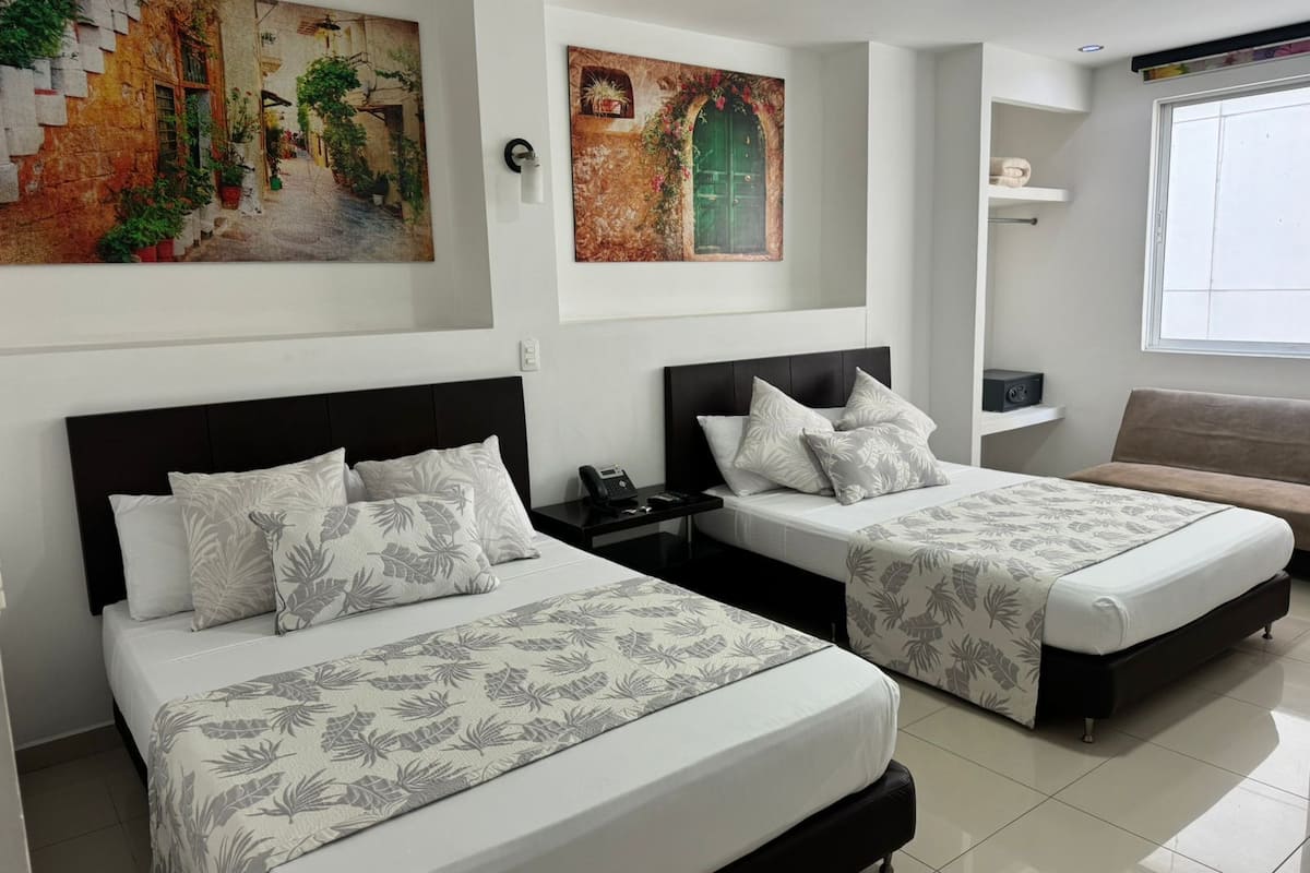 A hotel room with two beds, each with a black headboard and floral-patterned bedding. There are wall-mounted paintings depicting outdoor scenes, a bedside table with a phone, and a window with a view of the outside.