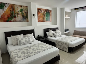 A hotel room with two beds, each with a black headboard and floral-patterned bedding. There are wall-mounted paintings depicting outdoor scenes, a bedside table with a phone, and a window with a view of the outside. - HGO HOTEL  (Cali)