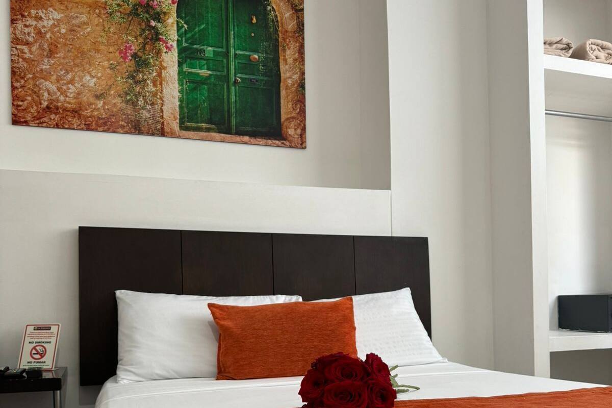 A neatly made bed with white and orange bedding, a green door with flowers, and a painting above it.