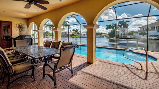 A poolside area with a table and chairs, a ceiling fan, and a view of the pool and surrounding buildings. - Heated Pool - Wide Gulf Access Canal - BBQ - Dock (Cape Coral)