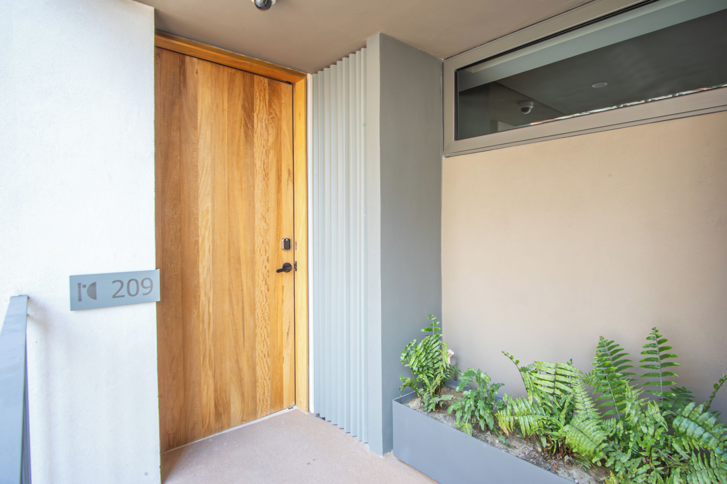 A modern entrance with a wooden door, a number sign, and a planter with ferns.