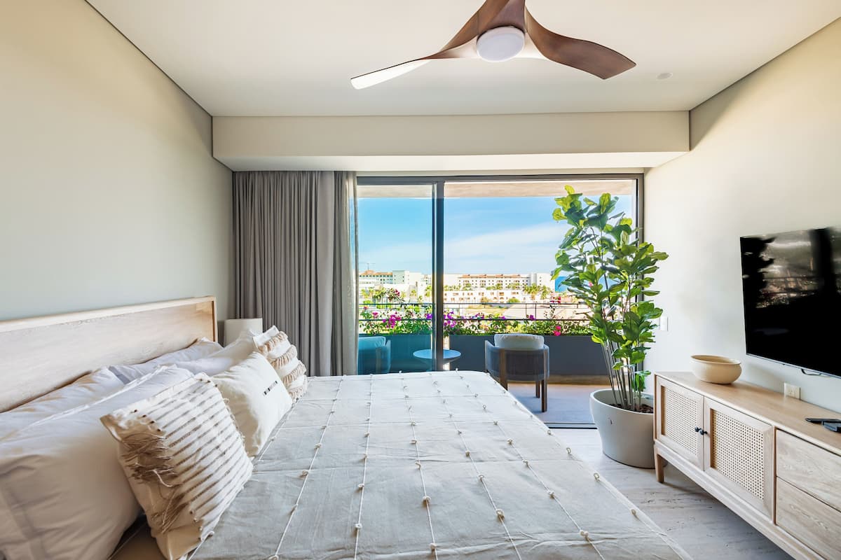 A modern bedroom with a large bed, a TV, a ceiling fan, and a balcony with a view of the city.