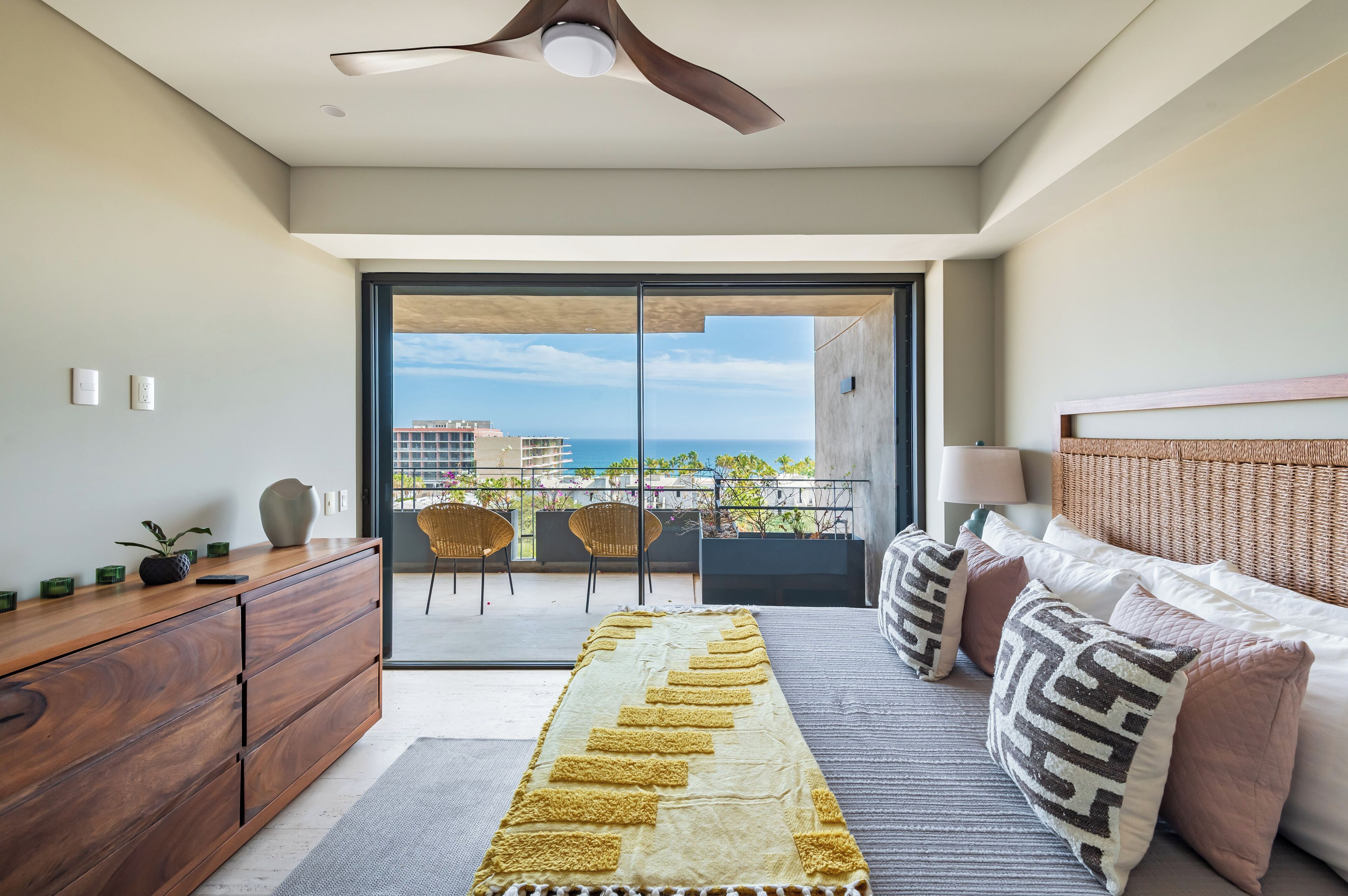 Une chambre moderne avec balcon, une commode en bois, un lit avec des oreillers et une vue sur l’océan et les bâtiments.
