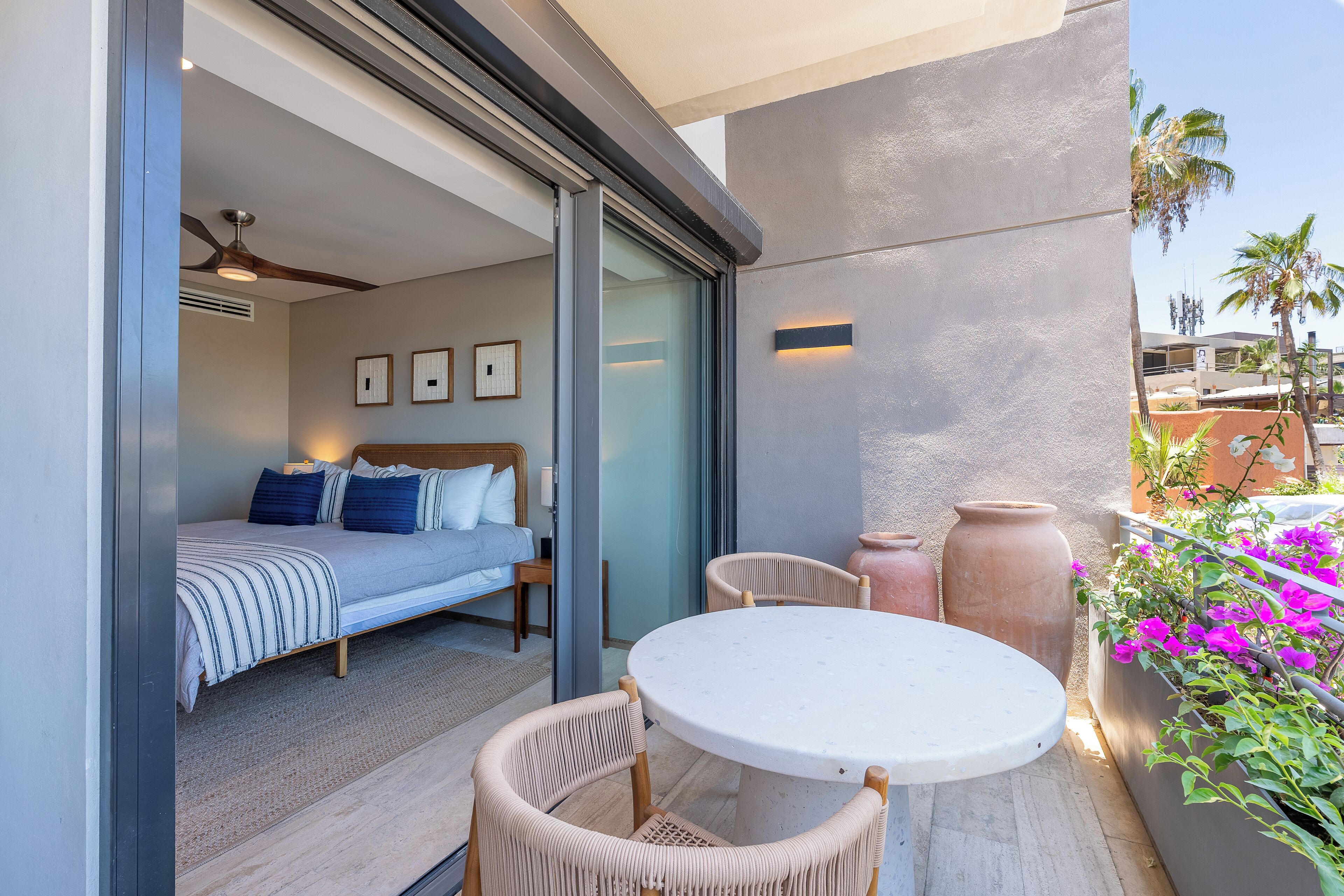 A modern bedroom with a bed, bedside table, and a ceiling fan. There is a balcony with a table and chairs, and a large planter with flowers.