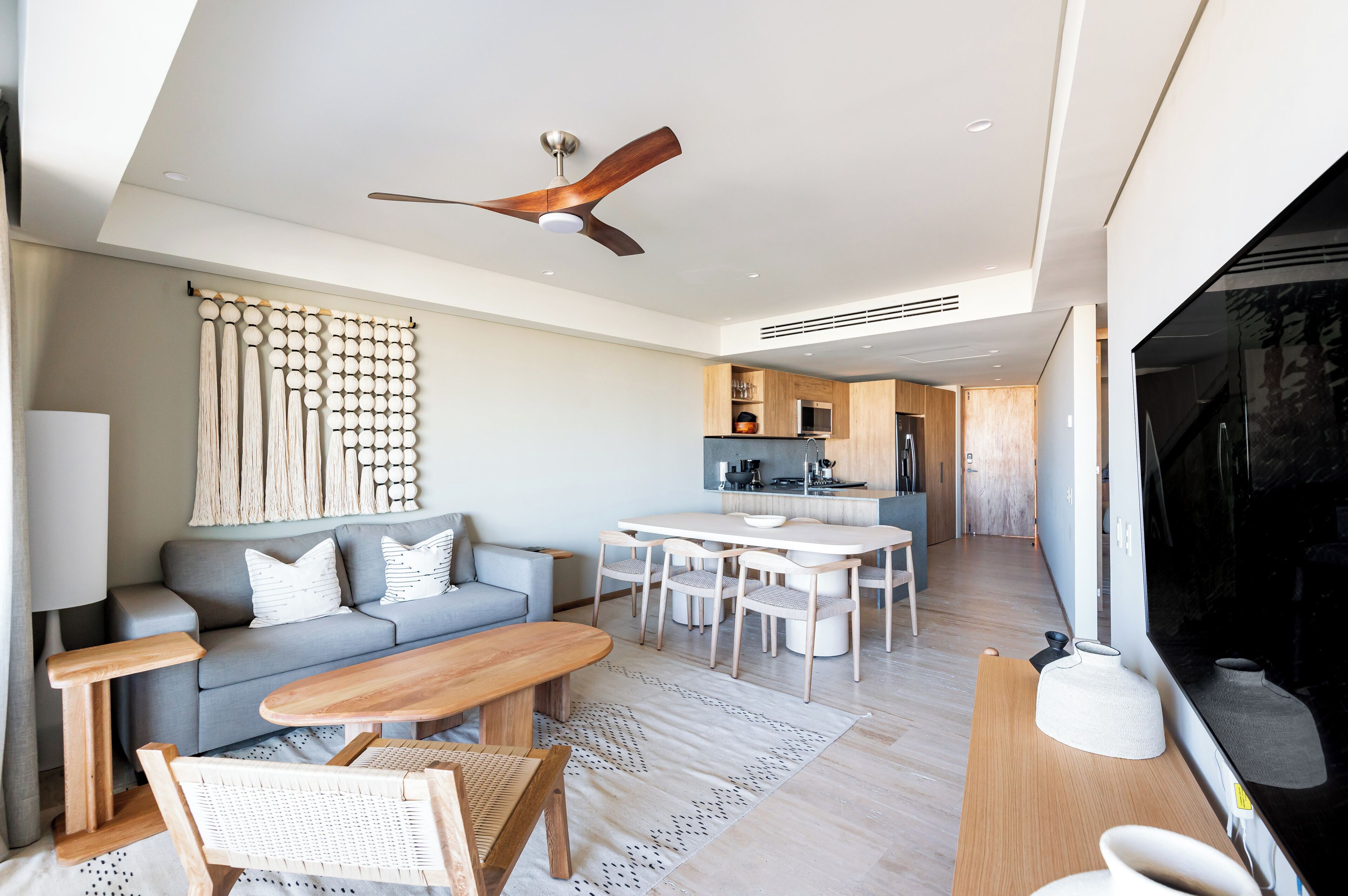 A modern living room with a grey sofa, wooden coffee table, and a dining area with a white table and chairs. A ceiling fan is visible, and there is a kitchen area in the background.